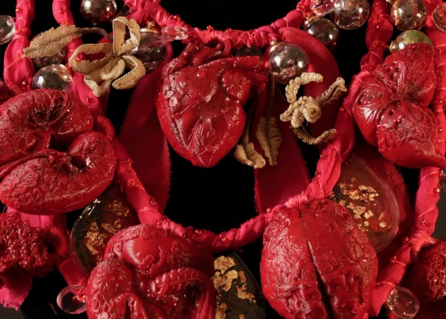 A vivid red wearable artwork made from fabric, sculptural organ-like forms, beads and metallic details