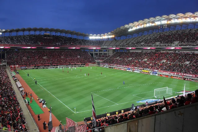 A packed soccer stadium lit under floodlights at night, stands full of spectators
