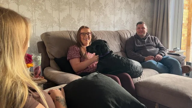 three people in a living room, a woman on a sofa holding a black dog, a man beside her, and a young woman in the foreground