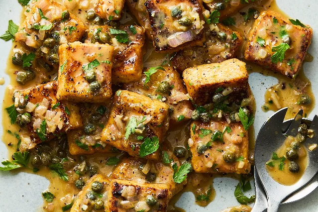 Golden pan-fried tofu pieces in a pale lemon-caper butter sauce with green capers, diced shallot and parsley leaves, in a pale ceramic dish with a serving spoon at the right edge