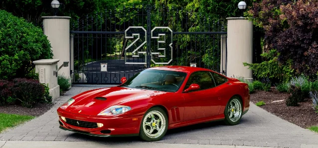 A red Ferrari 550 Maranello parked in front of tall gates bearing a large number 23