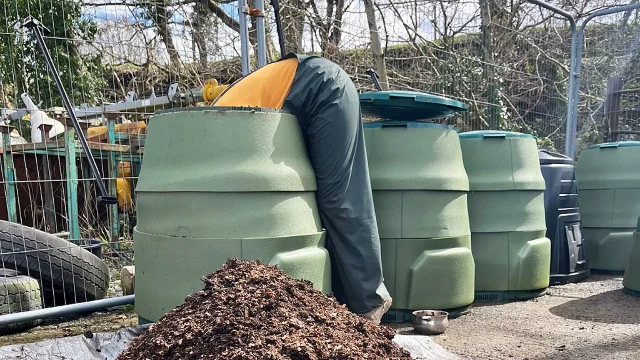 A worker leans into a green composting bin beside a mound of finished material.