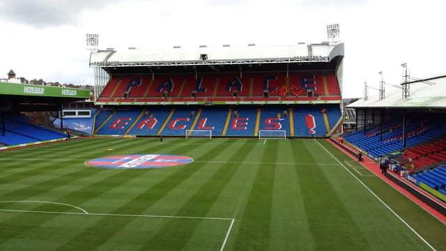 "A football stadium stand with blue seating, seen across the pitch from an upper corner of the ground"