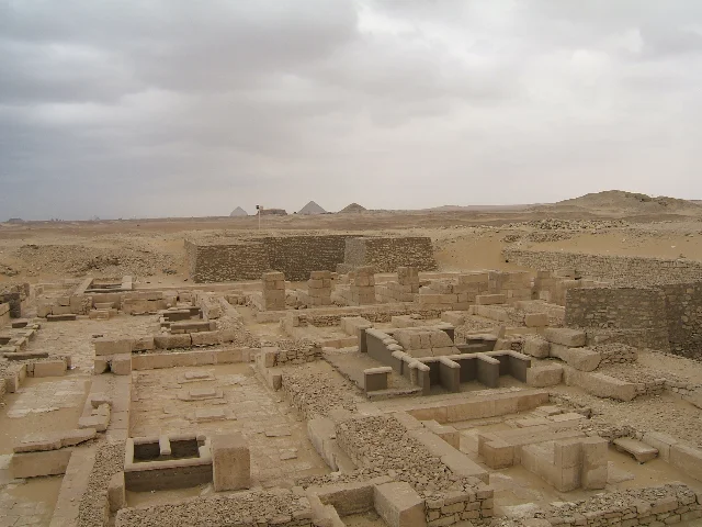 Excavated stone foundations and low walls in sandy desert, with three pyramids on the horizon under a grey sky