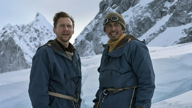 Two climbers in blue parkas roped together on a glacier below snow-covered peaks