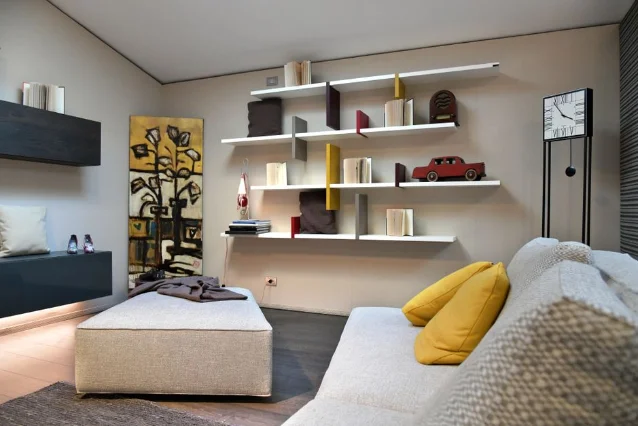 Multiple white floating shelves with colorful books and small decor objects mounted on a living room wall