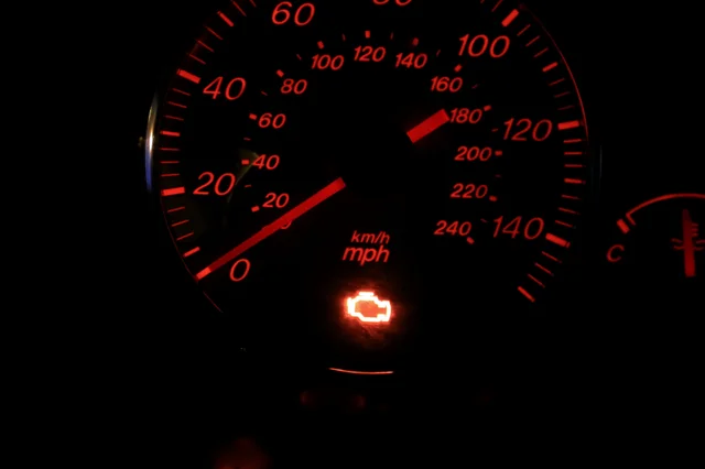 The instrument cluster of a car at night, speedometer and tachometer lit in red, with the amber engine-shaped warning symbol glowing beneath the dials