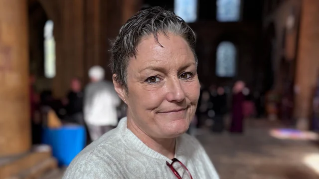 a woman with short grey hair in a pale jumper, smiling at the camera inside a large stone building
