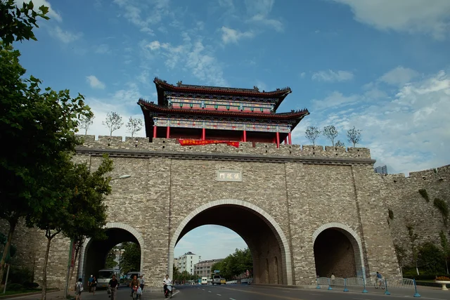 A three-arched stone gate in a long grey brick city wall, with a red pavilion on top