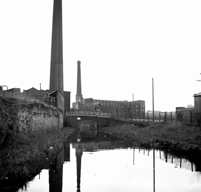 A black-and-white photograph of a narrow canal running between stone walls, with two tall mill chimneys and a mill building behind a bridge