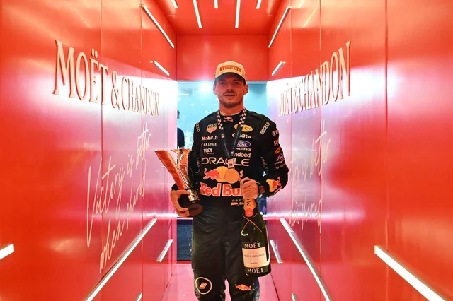 Max Verstappen in Red Bull race suit holding a trophy and a bottle of champagne in a red corridor