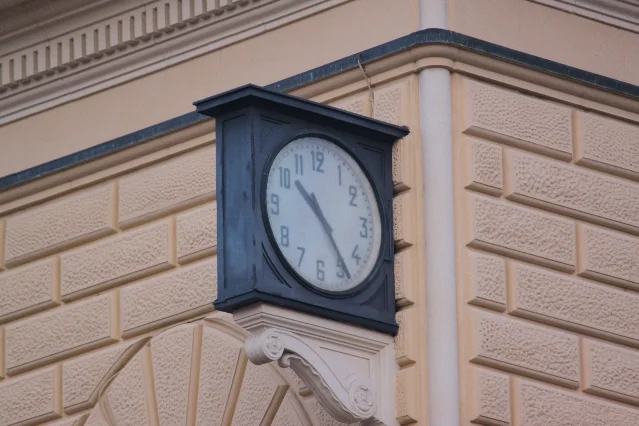 A dark-cased station clock with a white face and black hands, mounted on the corner of a pale stone building