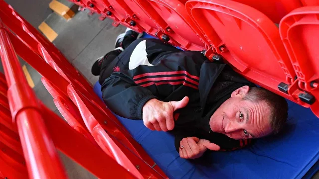 Chris Kirkland lying on a blue mattress beneath rows of red stadium seats, giving a thumbs-up