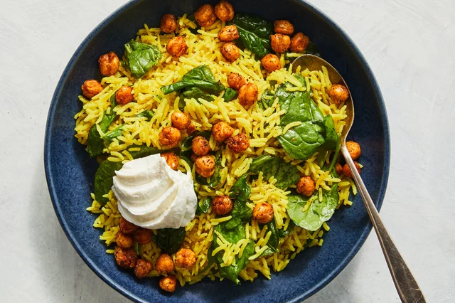 A deep blue bowl of golden turmeric rice topped with rust-red harissa-roasted chickpeas, wilted spinach leaves, and a dollop of white yogurt, with a bronze spoon resting in the bowl