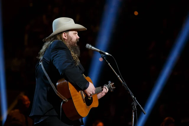 A bearded man in a pale cowboy hat and dark suit playing an acoustic guitar at a microphone
