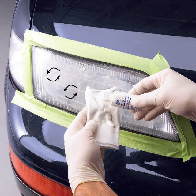 A masked headlight lens on a dark car with two printed circular arrows on the plastic, as gloved hands hold a small white tube of plastic polish and a folded polishing cloth