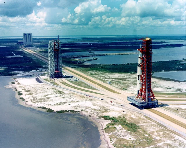 An aerial view of a Saturn V on its crawler moving along the crawlerway, with a second launcher and the assembly building in the distance