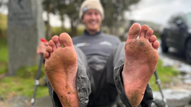 a man sitting on a wall holding up two bare feet covered in gravel and mud, his face out of focus behind them
