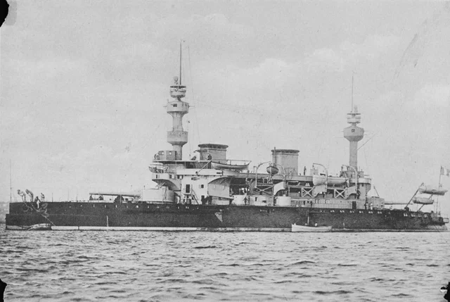 A broadside photograph of a battleship at anchor, with two tall masts, boats stowed high on the superstructure and a dark hull