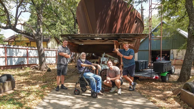 four men in front of a large steel boat hull in a residential yard, one of them seated on a wheeled walker