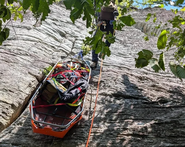 An orange metal rescue litter rigged with ropes and gear rests on a steep rock face as a rescuer ascends above in Linville Gorge.