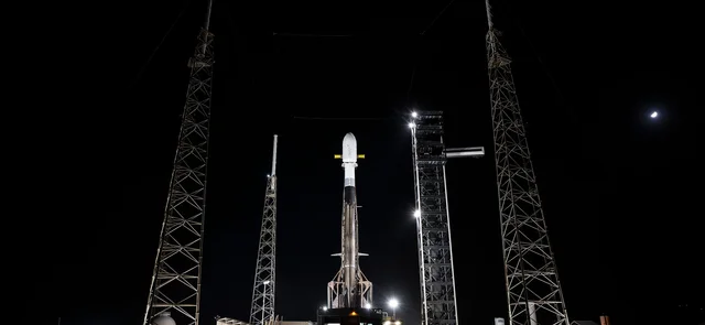 A SpaceX Falcon 9 with its payload fairing stands upright on Space Launch Complex 40 at night, lit by pad spotlights between lightning towers, with the Moon visible at upper right