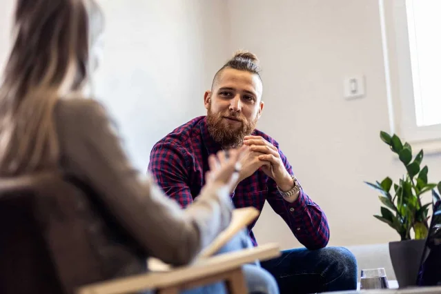 Two people in conversation across a small table in a bright room, a plant beside them.