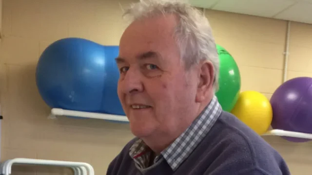 A grey-haired man in a checked shirt and dark jumper, with coloured exercise balls on a shelf behind him