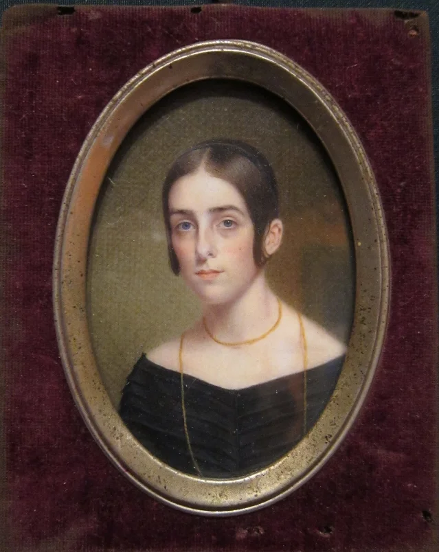 An oval portrait of a young woman in a black dress and gold chain, in a metal frame on dark velvet