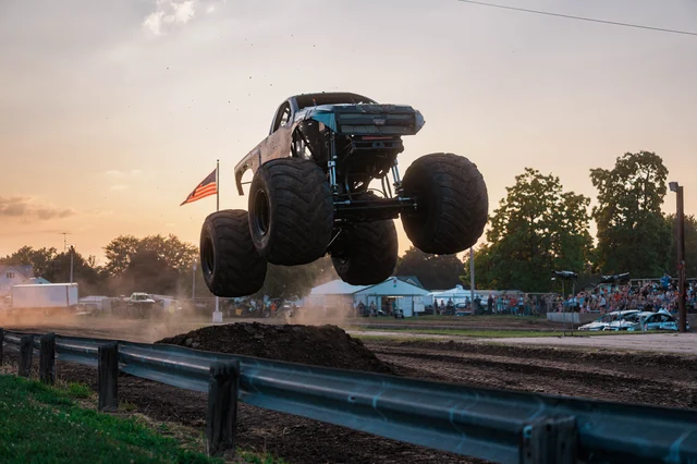 A monster truck airborne over a dirt mound at sunset, with a crowd and an American flag behind