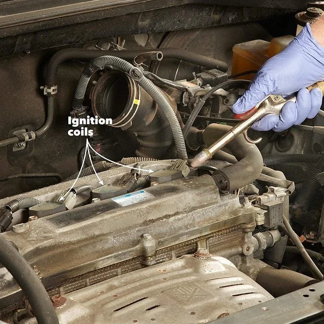 A gloved hand uses a ratchet beside ignition coils in an engine bay.