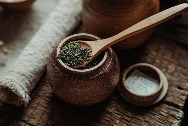 Loose green tea held in an open ceramic jar with a wooden spoon, its lid set beside it