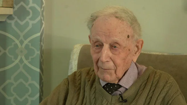 An older man in a green cable-knit jumper and a striped shirt, seated in an armchair