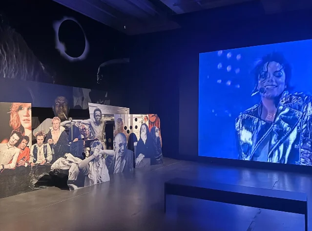 展厅里正前方一块屏幕上是一位闭眼张口唱歌的女性黑白特写，左侧墙上是几名歌手的剪影拼贴