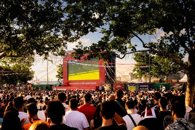A large outdoor fanzone crowd watches a live soccer match on a big screen under warm evening light