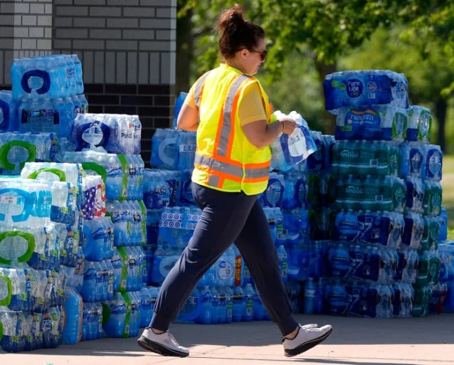 A volunteer in a safety vest carries water past stacks of donated bottles.