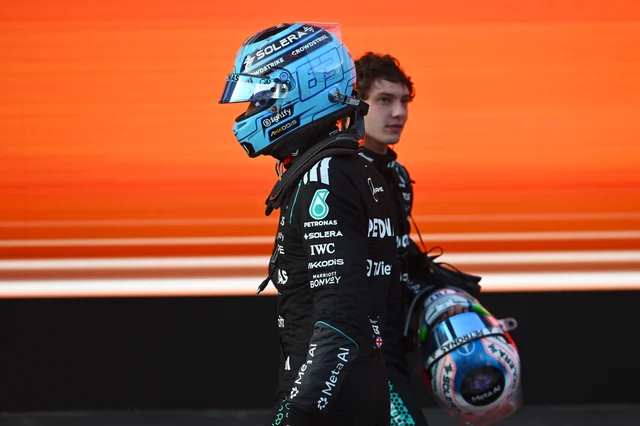 Two Mercedes Formula 1 drivers in black race suits carrying helmets, walking in front of an orange barrier