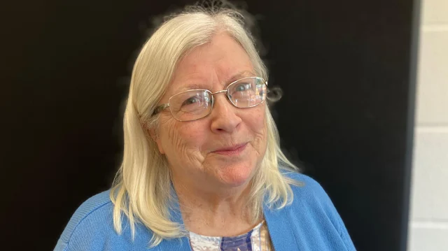 Pat, 77, wearing glasses and a blue cardigan, photographed indoors against a dark wall