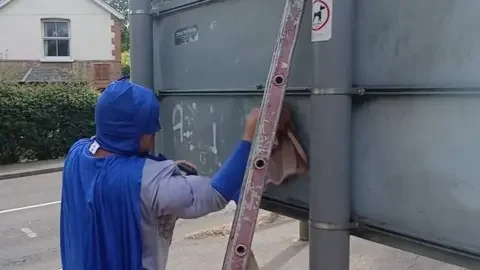 Liam Keen dressed as Batman cleans graffiti from a street sign.