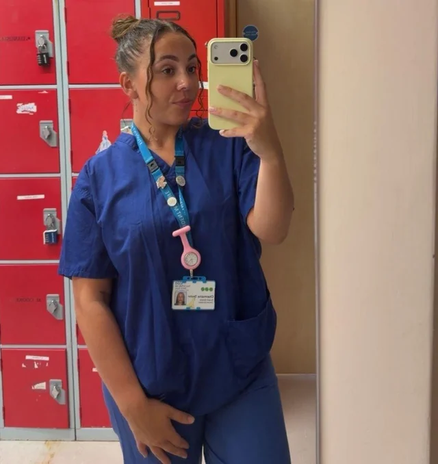 a woman in blue scrubs holding a phone up to a mirror in a hospital changing room, red lockers behind her