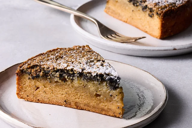 A wedge of pale golden cake on a small ceramic plate, with a dark black sesame layer running under a crackled, sugar-dusted top and a fork resting on a larger plate of cake behind it