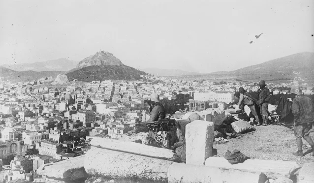 Soldiers at a machine-gun post on high ground, looking out over a dense white city with a rocky hill beyond