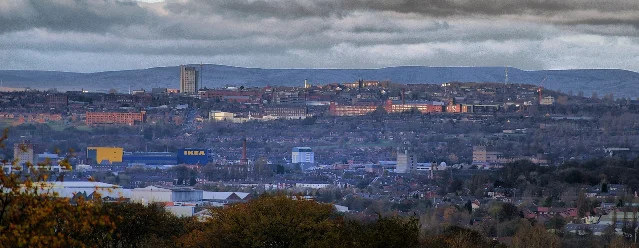 A wide view of a town filling a valley below a ridge of hills, with terraced streets, tower blocks, a brick chimney and a large blue and yellow store