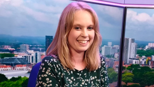 A smiling young woman with long blonde hair in a dark floral top, seated in front of a studio backdrop
