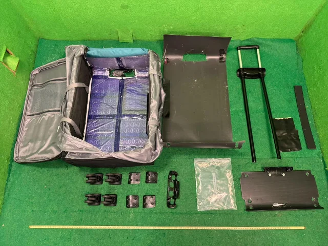 An opened grey suitcase on a green inspection floor with its false base fully disassembled — blue-wrapped parcels visible in the hollow cavity, and the metal false-bottom panel, trolley mechanism, and hardware laid out beside it