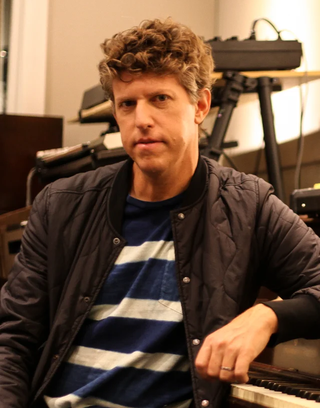 A man in a dark jacket and striped shirt sitting in a studio in front of keyboards