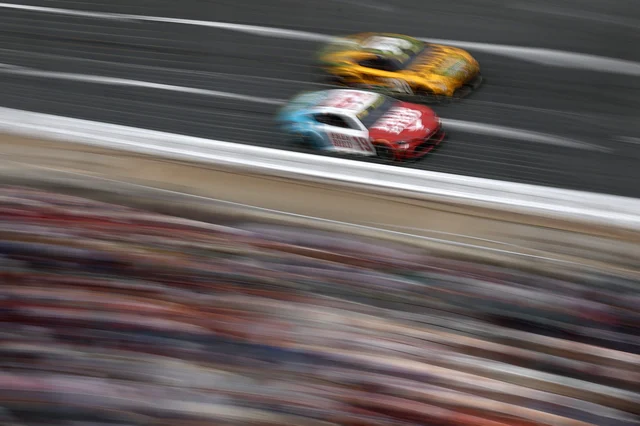 Two NASCAR Cup cars blurred by speed, racing side by side with a crowd in the foreground