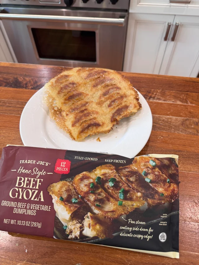 A white plate on a wooden table holding six pan-fried beef dumplings joined into one continuous golden-brown crisp sheet, flipped crisp side up, with a bite taken out of one edge; the maroon Trader Joe's bag lies in front of the plate and a stainless steel oven fills the background