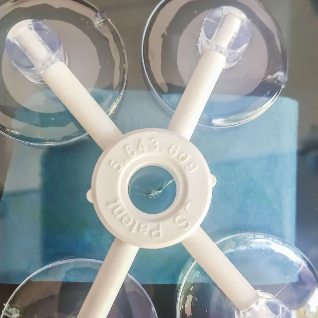 A white cross-shaped windshield repair fixture with four clear suction cups pressed onto glass, its centre ring sitting exactly over a small chip visible through the hole