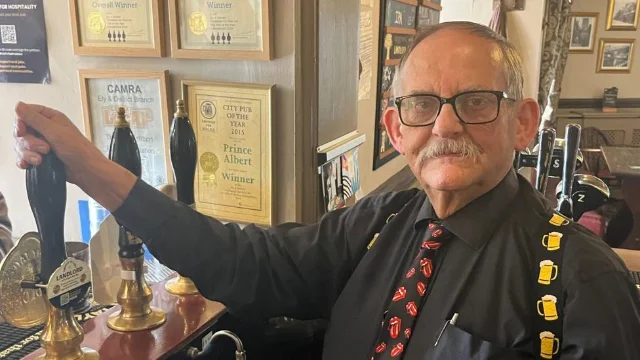 An older man with glasses and a moustache in a black shirt, novelty tie and beer-mug braces, pulling a beer tap behind a pub bar with framed certificates on the wall behind him